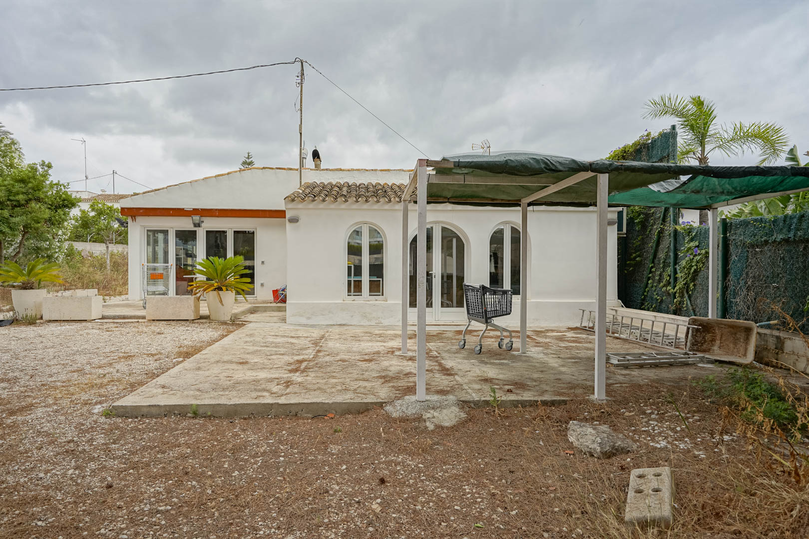 Exterior de la villa de una planta con ventanas de arco, zona de grava y una estructura de sombra metálica sobre base de hormigón.