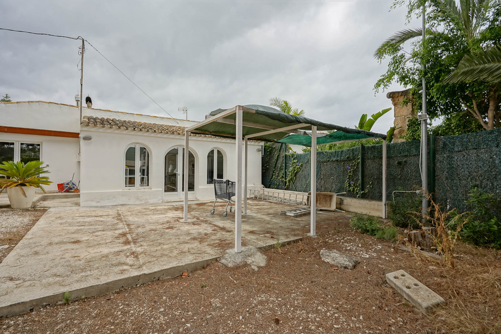 Exterior de la vivienda actual con fachada blanca y ventanales de arco, destacando un patio de hormigón y una estructura de pérgola junto al jardín perimetral.