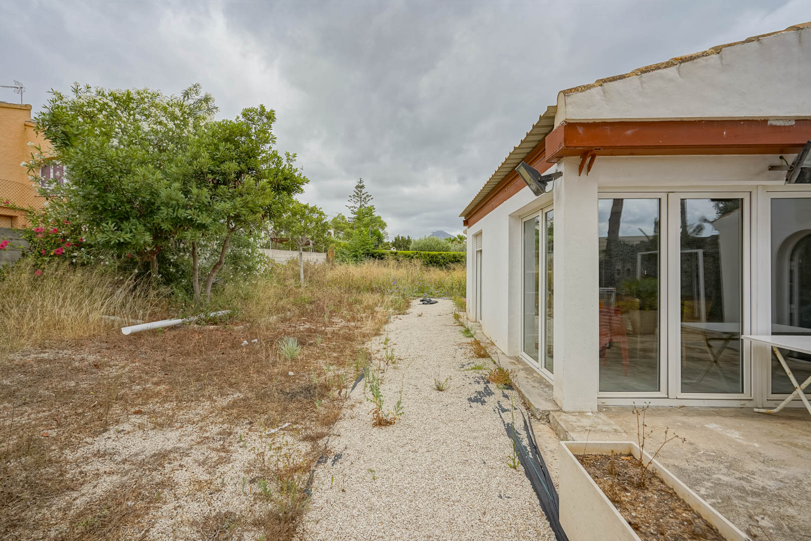 Fachada lateral de la vivienda con grandes ventanales de PVC blanco y acceso a una amplia parcela de terreno natural con vegetación mediterránea.