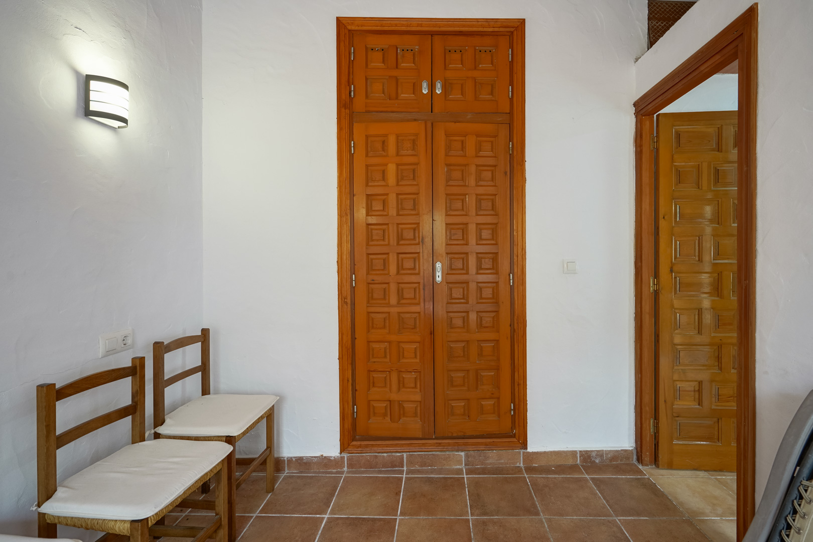 Pasillo con suelos de gres rústico, paredes blancas y armarios empotrados de madera tallada. Incluye iluminación de pared y acceso a estancias contiguas.