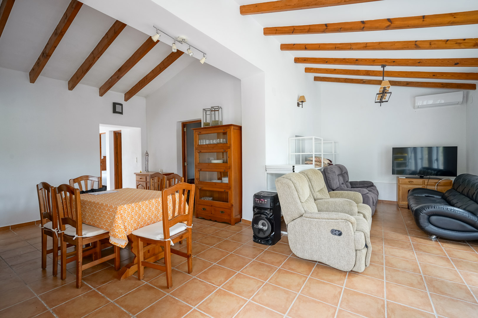 Salón-comedor diáfano con techos de vigas de madera vistas, suelos de gres rústico y mobiliario clásico de madera en un ambiente luminoso.