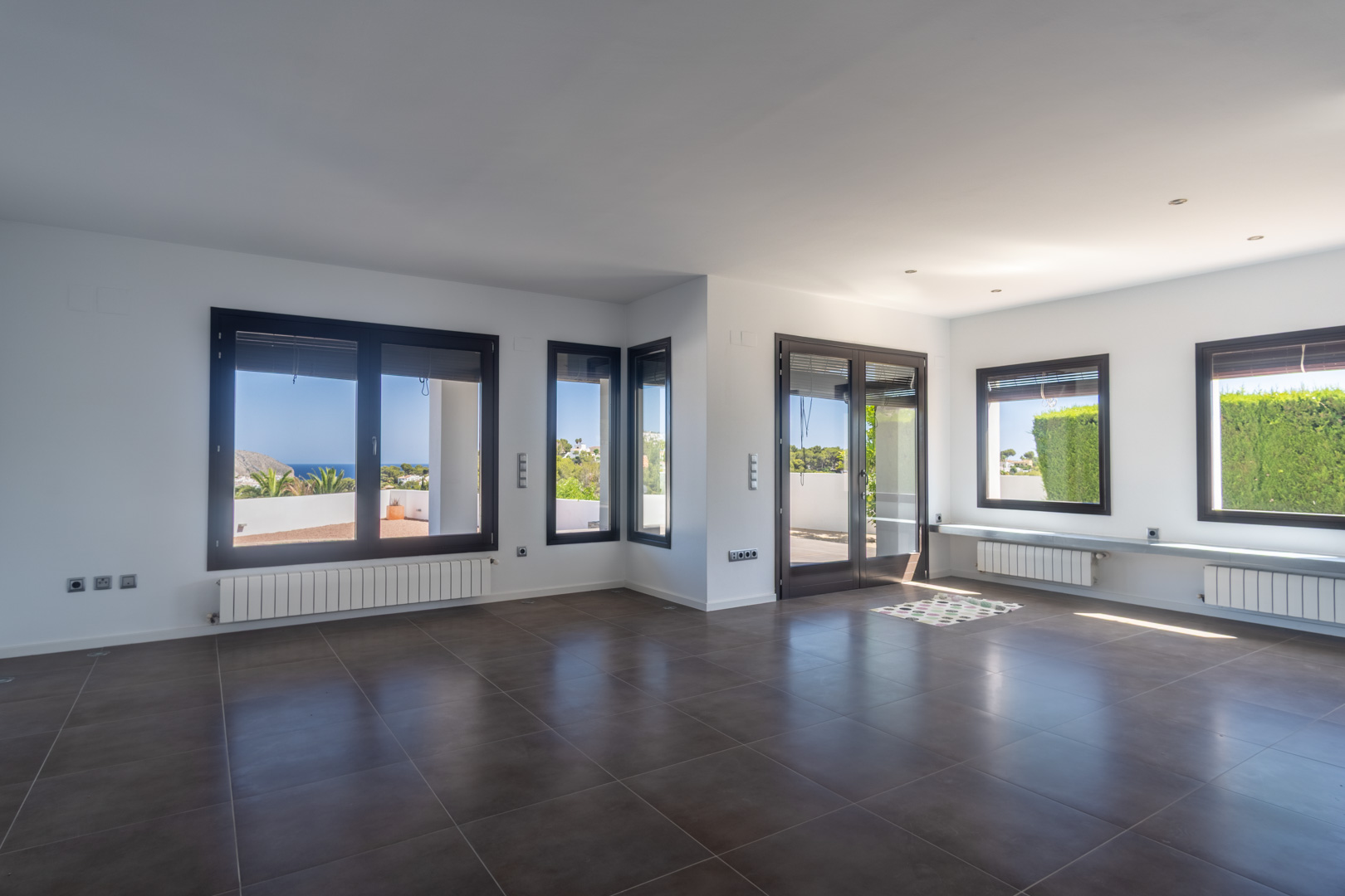 Amplio salón comedor diáfano con suelo cerámico oscuro, radiadores bajo ventanales de aluminio efecto madera y vistas panorámicas al mar y la costa.