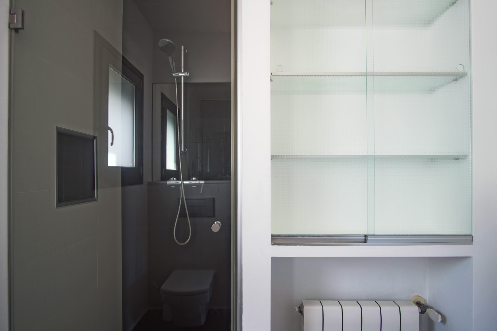 Baño moderno con azulejos grises, ducha con grifería cromada y sanitario suspendido, junto a un armario empotrado con estantes de cristal y radiador.