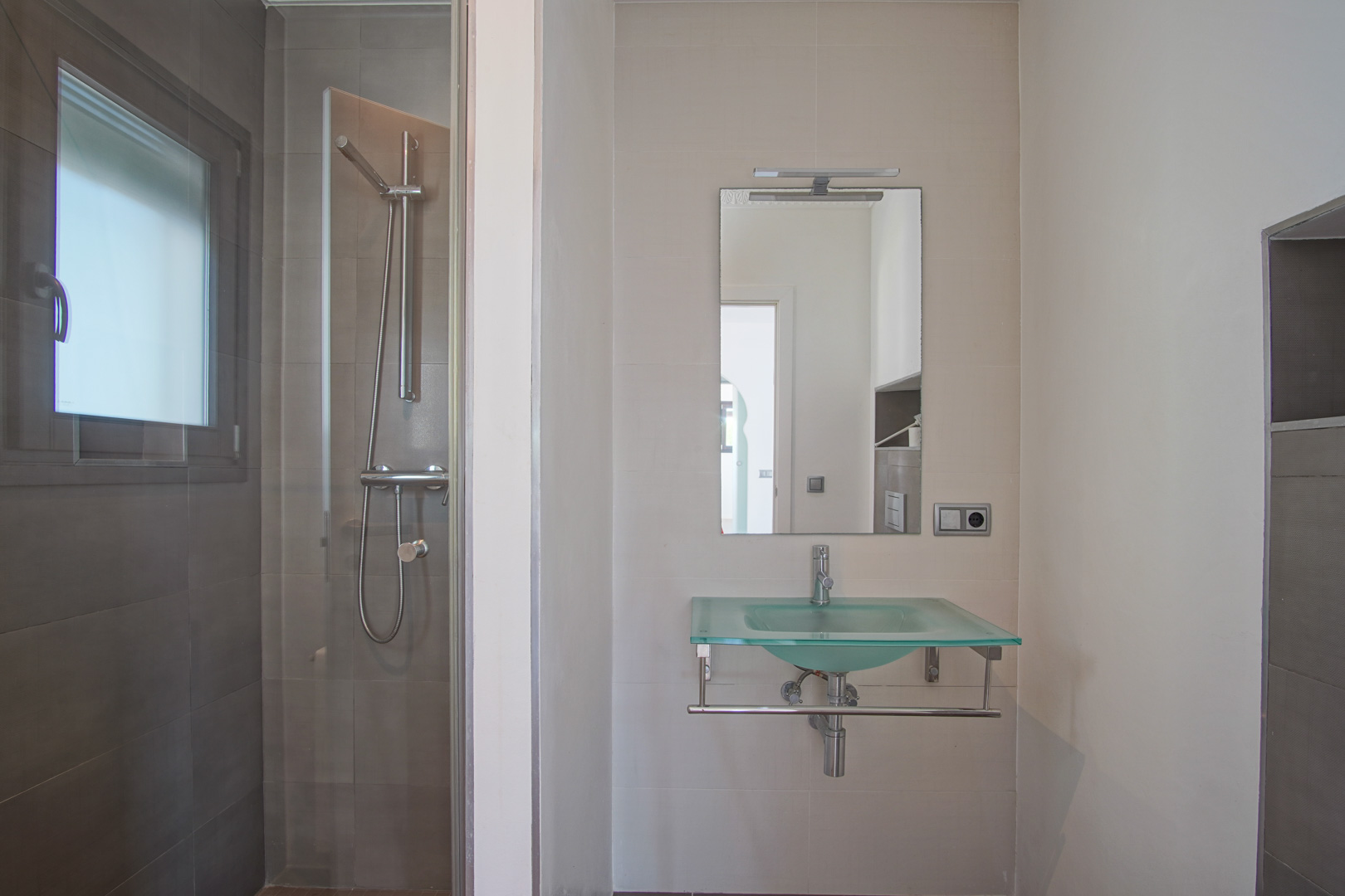 Baño moderno con lavabo de cristal translúcido, espejo con iluminación superior y ducha de obra con mampara de cristal y azulejos en tonos grises.