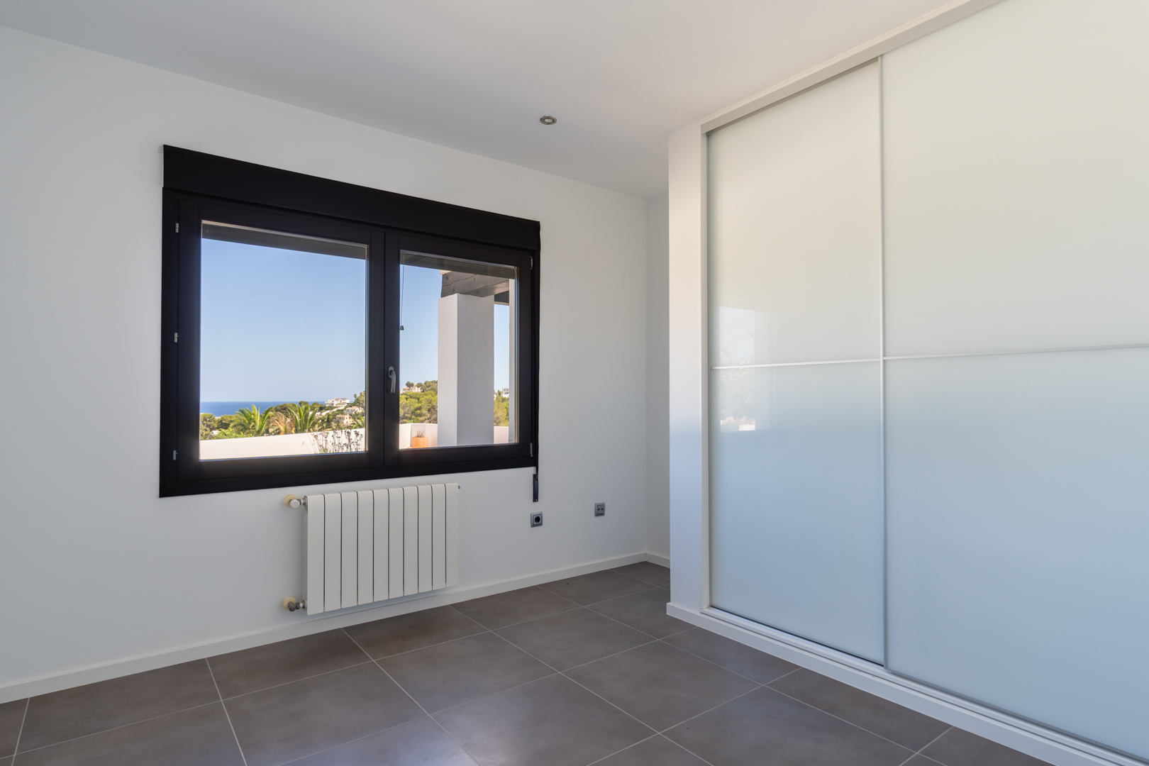 Dormitorio luminoso con armario empotrado de puertas correderas, radiador y ventana de marco oscuro con vistas despejadas al mar y la costa de Moraira.