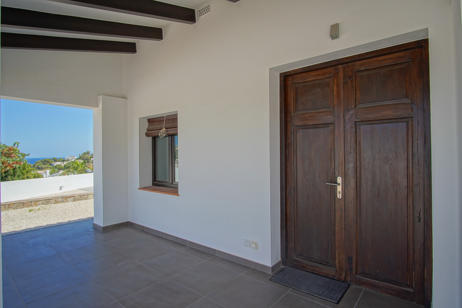 Entrada principal con puerta de madera antigua restaurada, vigas oscuras en el techo y porche cubierto con vistas al entorno costero de Moraira.