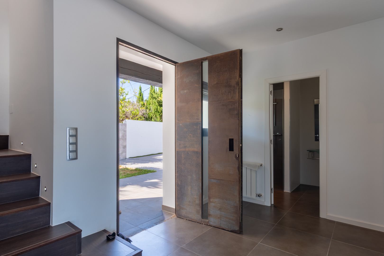 Entrada principal con puerta pivotante de efecto acero corten y cristal. Dispone de suelos cerámicos oscuros, escalera con iluminación LED integrada y acceso a un baño completo.