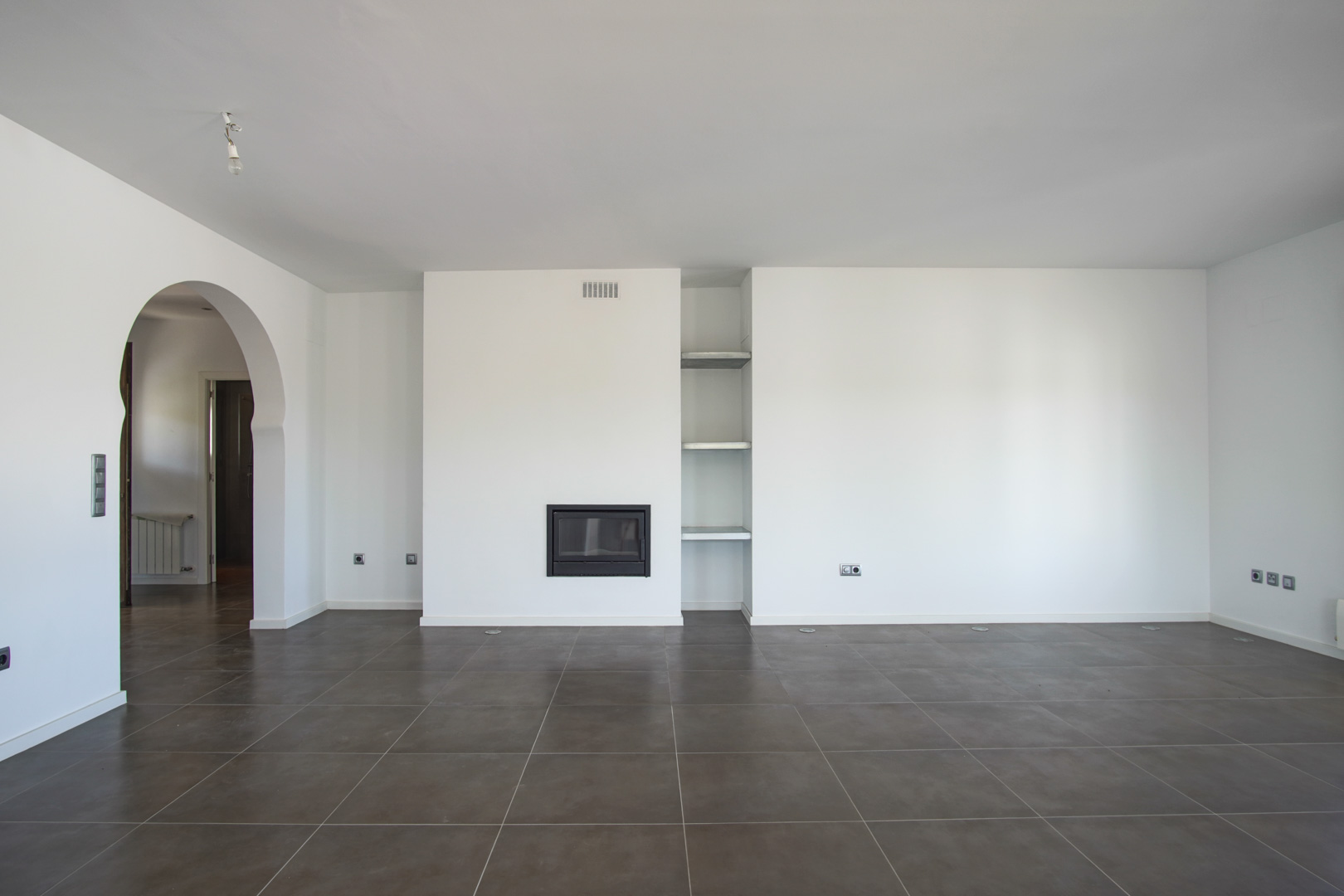 Salón diáfano con chimenea encastrada, estanterías de obra y suelos de gres oscuro. Un arco de estilo mediterráneo conecta con el pasillo y las estancias contiguas.