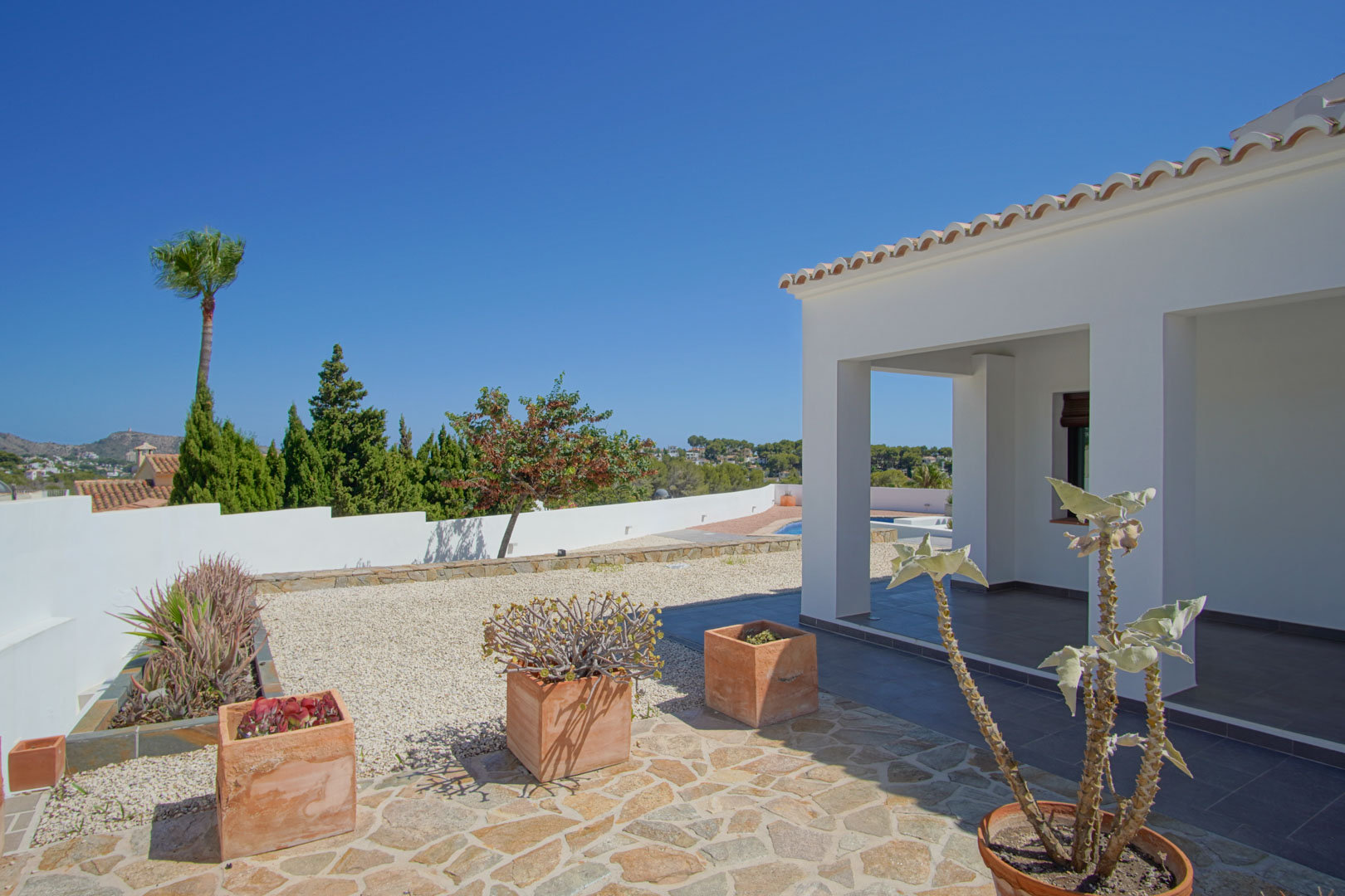 Terraza exterior con porche cubierto, pavimentos de piedra natural y maceteros de barro. El diseño integra vegetación mediterránea y ofrece vistas despejadas al entorno.