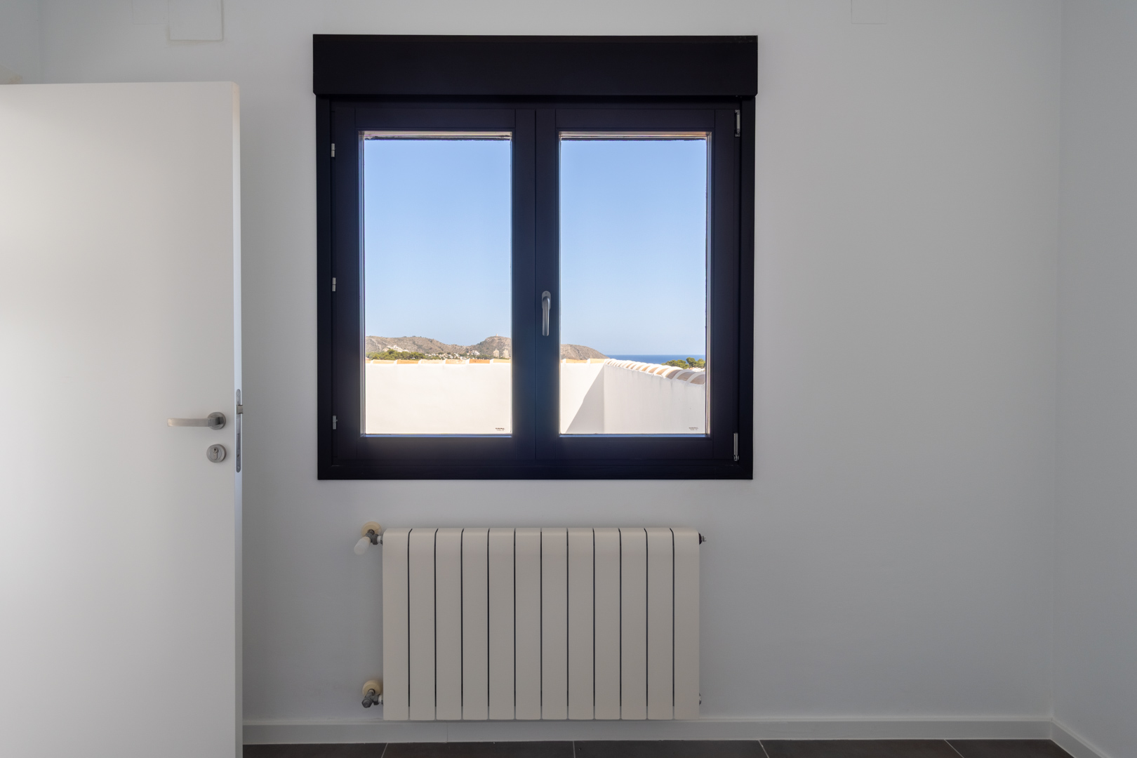 Ventana de aluminio oscuro con doble acristalamiento que ofrece vistas al mar y al Cabo de Moraira, situada sobre un radiador de calefacción en una estancia de paredes blancas.
