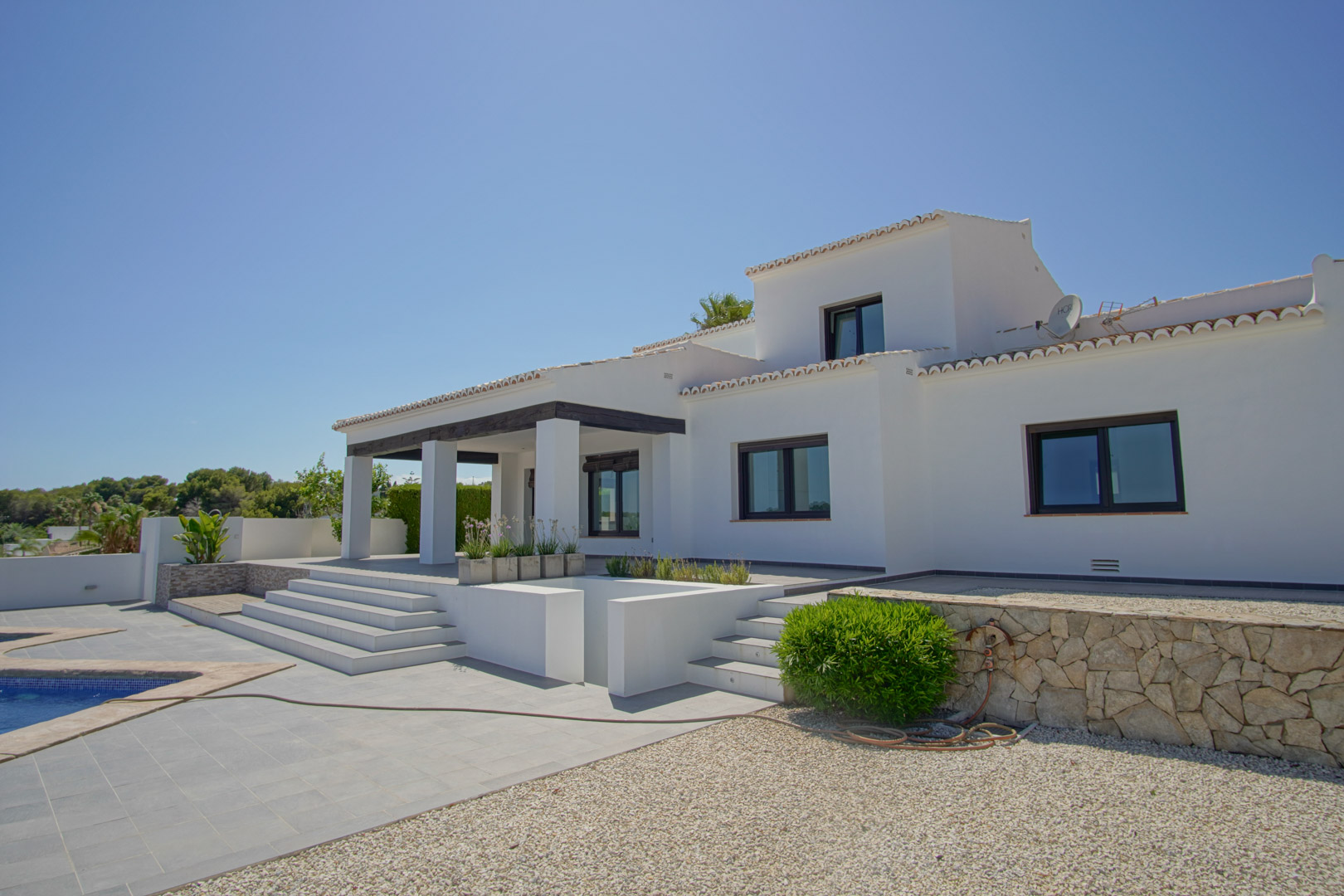Vista exterior de la villa mediterránea con fachada blanca, porche de vigas oscuras y terraza nivelada que conecta con la zona de piscina y muros de piedra.