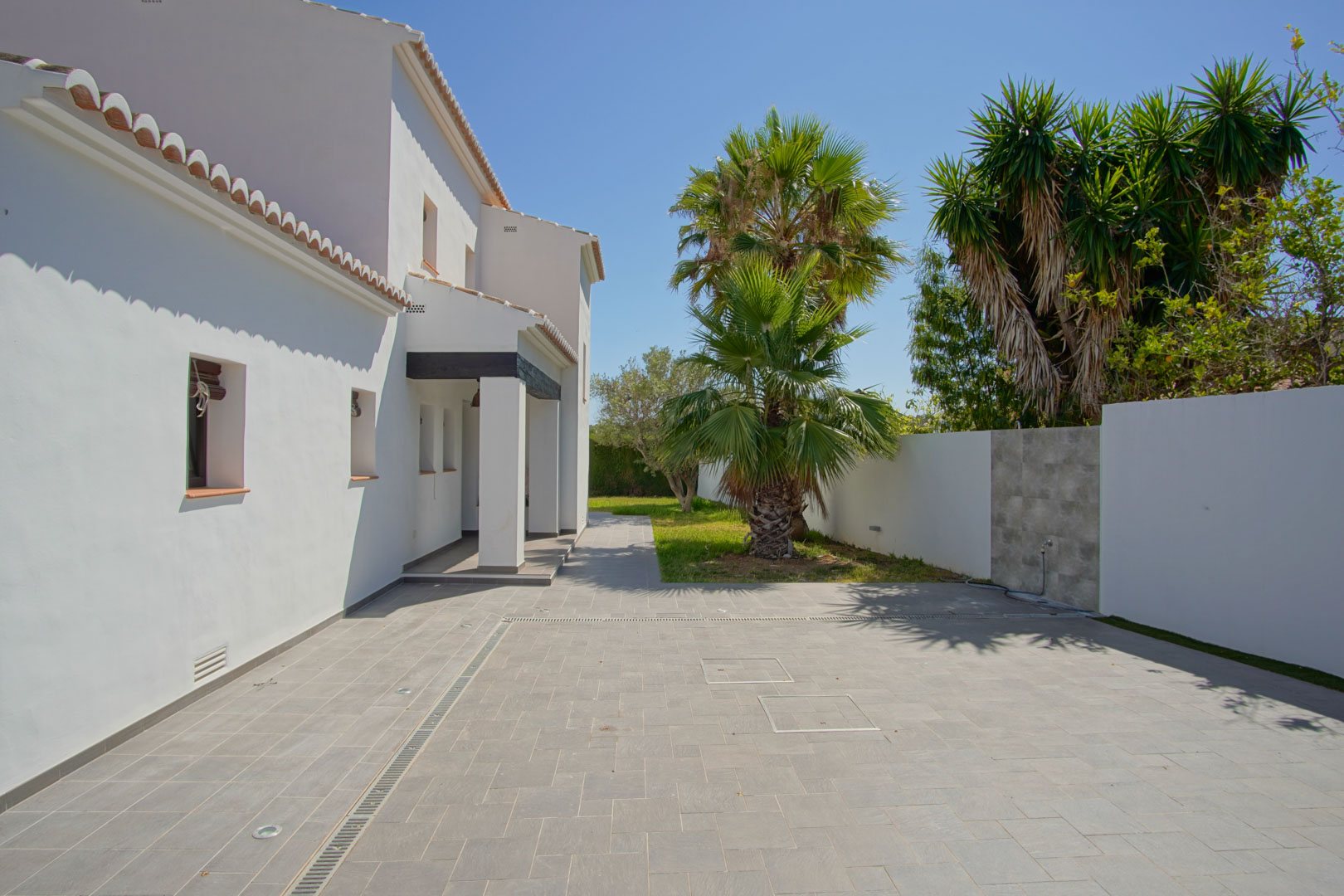 Zona de entrada pavimentada con gres gris, iluminación integrada en el suelo y jardín mediterráneo con palmeras junto a la fachada blanca de la villa.