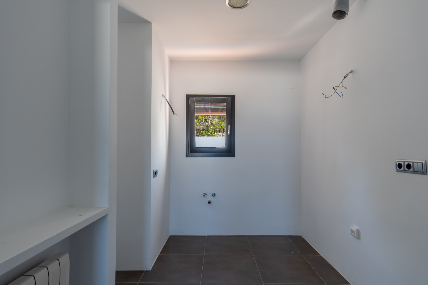 Zona de lavadero con preinstalaciones técnicas, suelos de gres oscuro y ventana de aluminio. Espacio funcional con acceso a tomas de agua y electricidad.