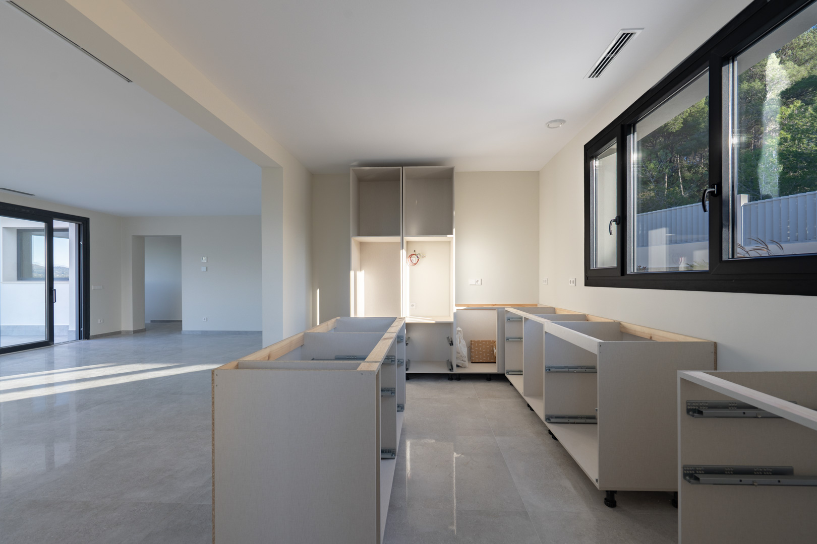 Cocina en obra con bases de armarios blancos y suelo de baldosa gris. Ventana con carpintería negra.