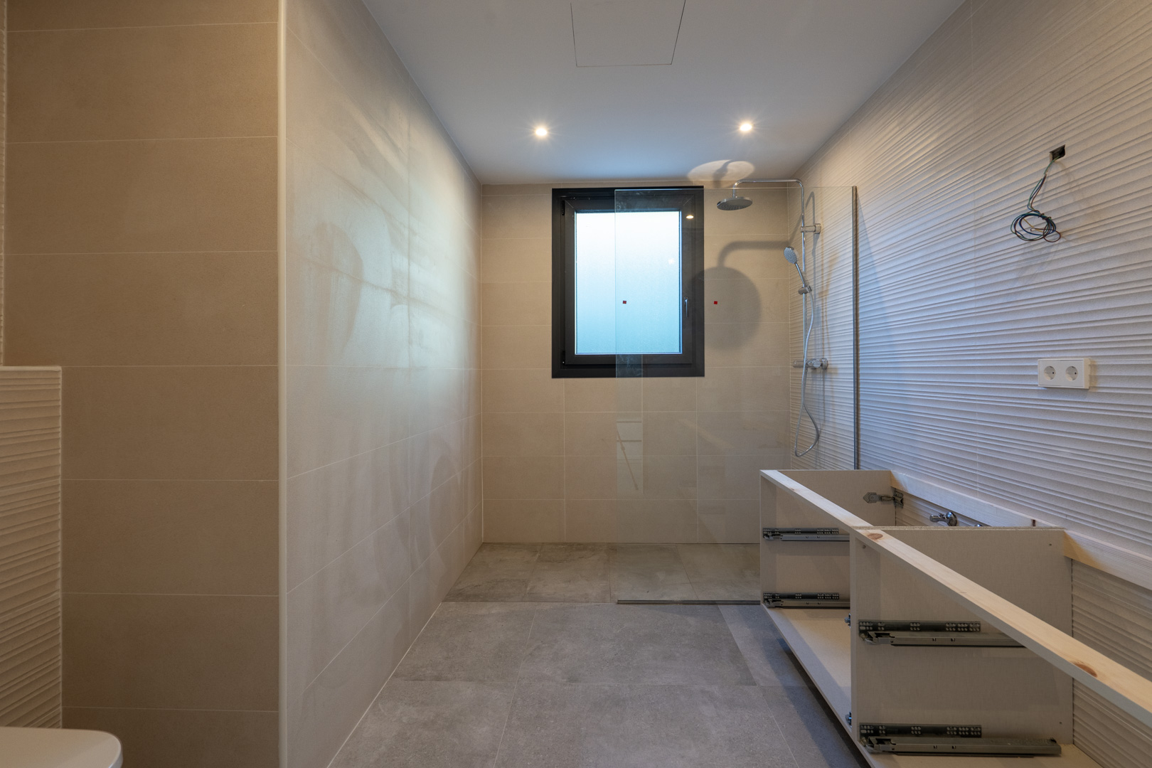 Detalle de baño moderno con ducha, ventana cuadrada y mueble de lavabo sin terminar.