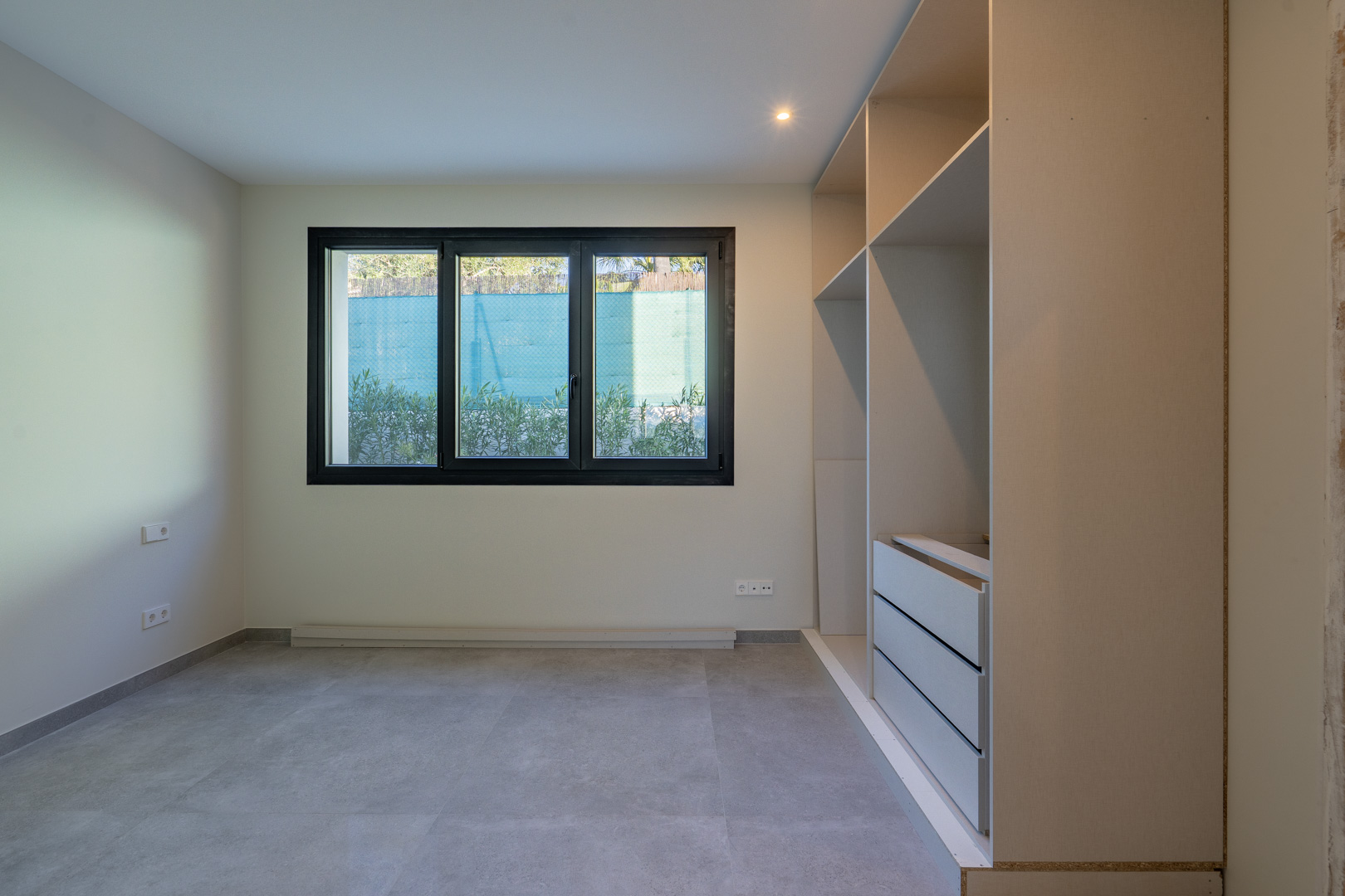 Dormitorio luminoso con ventana de triple panel y armario empotrado sin puertas. Suelo de baldosas grises.