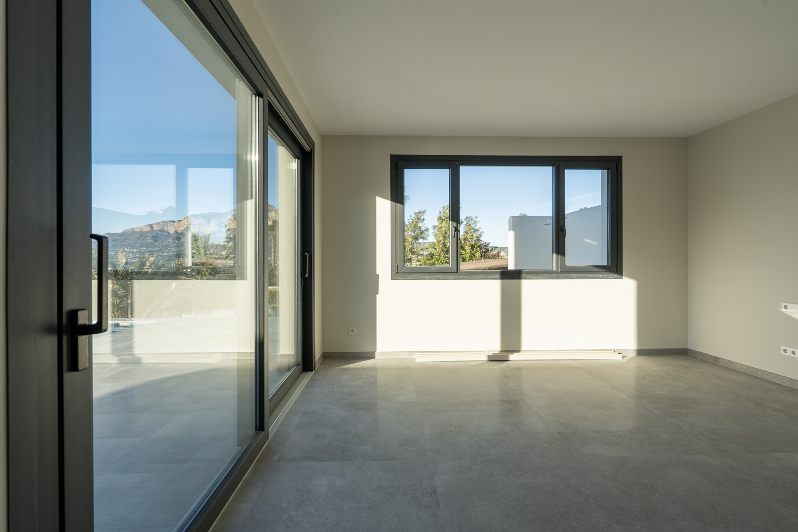 Luminoso espacio interior con suelo de baldosas grises, amplias ventanas correderas y vistas al exterior.