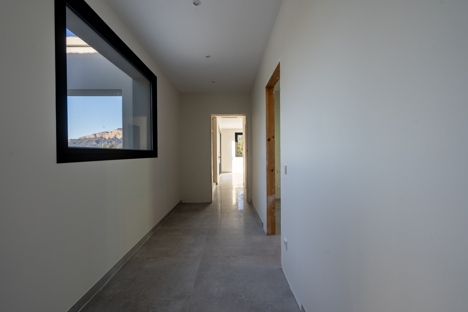 Pasillo luminoso con suelo de baldosas grises y vista a la montaña a través de una ventana rectangular con marco negro.