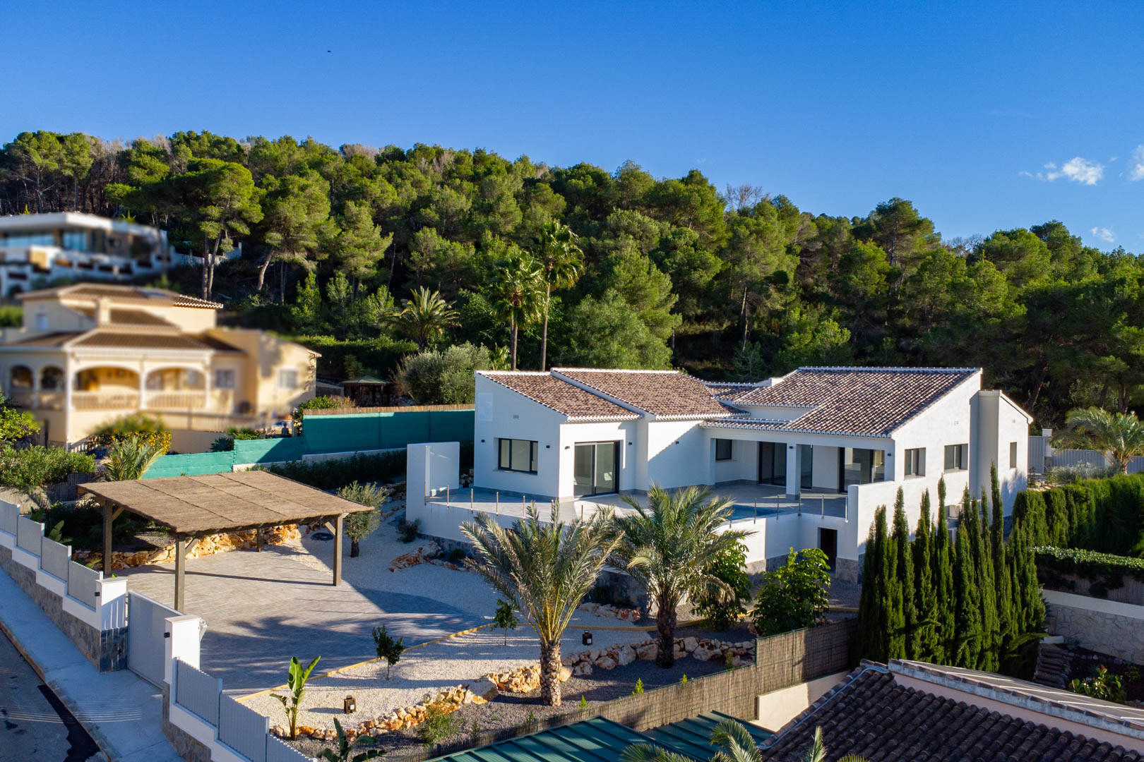 Vista aérea de villa moderna con tejas marrones, fachada blanca y carport techado en Jávea, rodeada de vegetación.