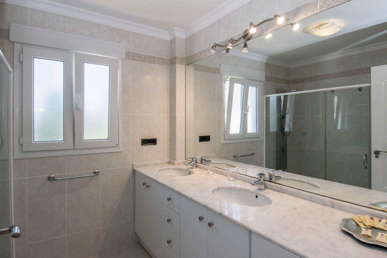 Cuarto de baño con encimera de mármol de doble seno, mueble blanco y gran espejo con iluminación integrada. Paredes alicatadas en tonos neutros y ventana oscilobatiente.