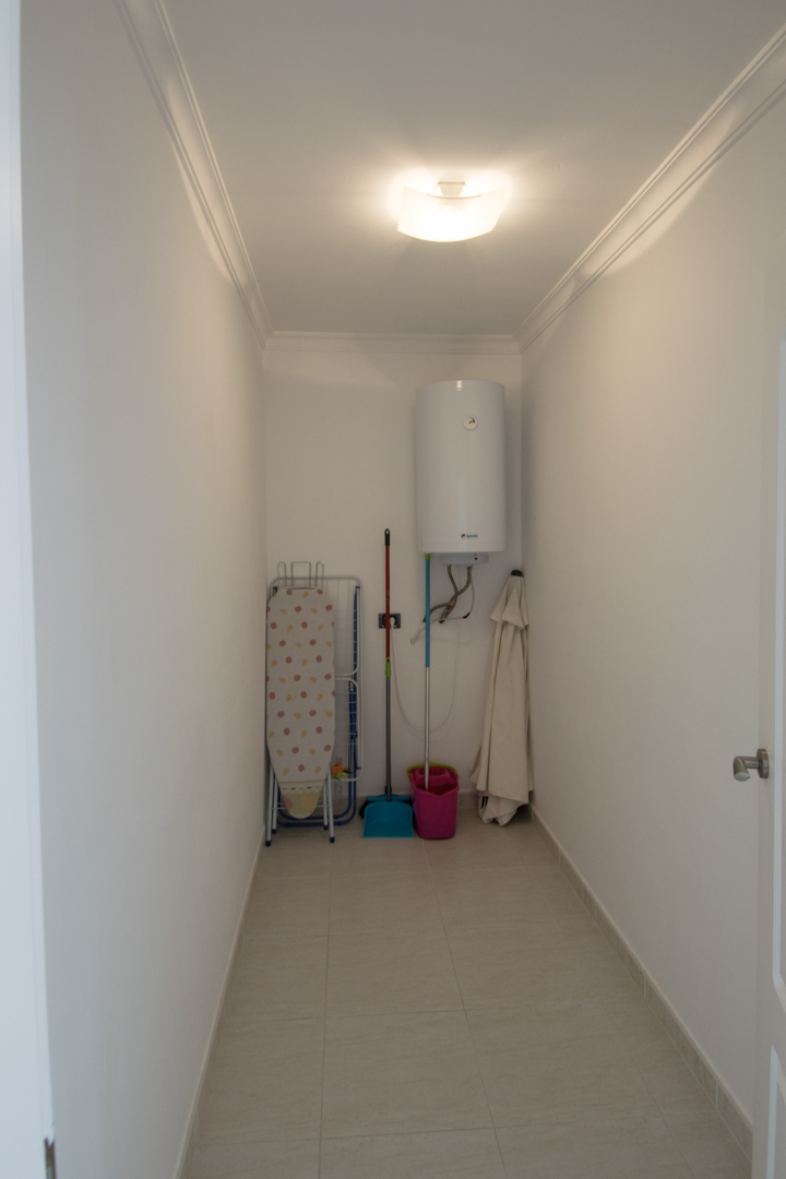 Cuarto de servicio funcional con termo eléctrico, espacio para artículos de limpieza y tabla de planchar, con suelos de gres y molduras.