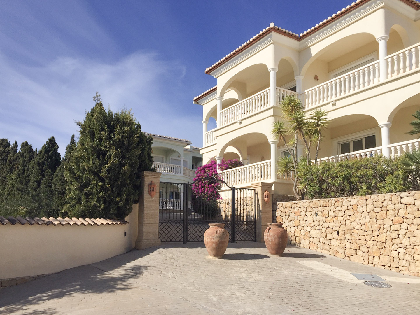 Entrada principal al complejo con puertas de hierro forjado, muros de piedra natural y edificios de estilo mediterráneo con amplias terrazas arqueadas.