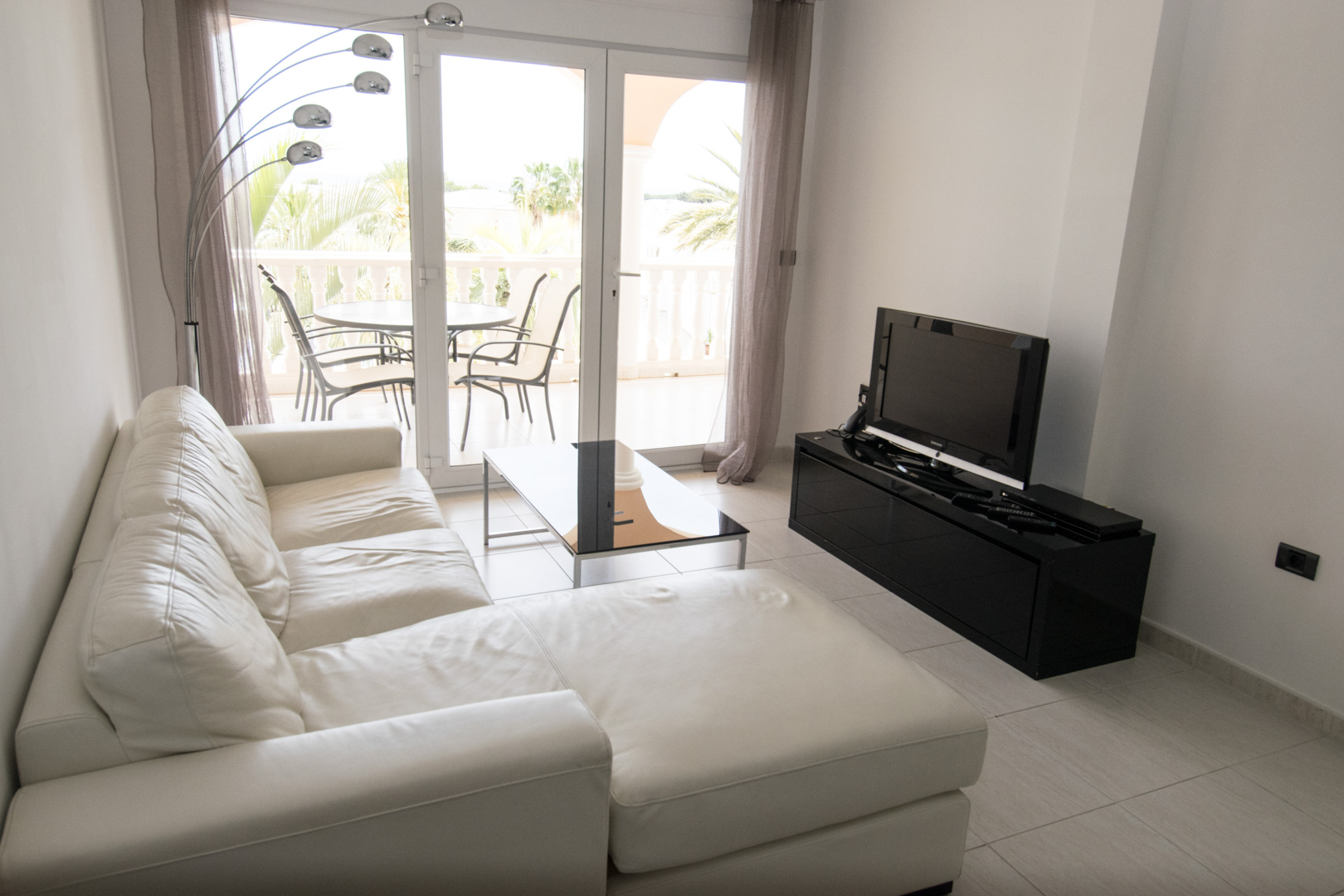 Salón luminoso con sofá de cuero blanco, mueble de TV negro brillante y acceso directo a una terraza privada con vistas a palmeras.