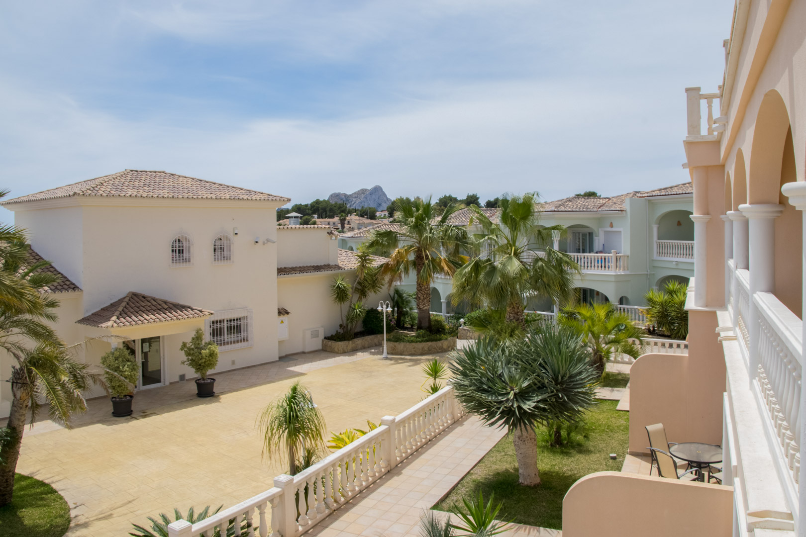 Vista desde la terraza del complejo que muestra la arquitectura mediterránea, jardines cuidados con palmeras y vistas lejanas al Peñón de Ifach bajo un cielo despejado.