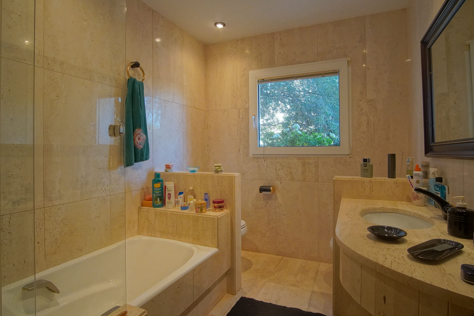 Baño revestido íntegramente en mármol crema con bañera, mampara de cristal y encimera de piedra natural. Incluye ventana exterior con vistas al jardín y luz empotrada.