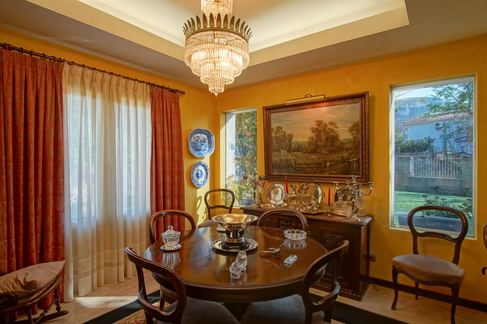 Comedor elegante con mesa circular de madera, lámpara de araña de cristal y aparador clásico. Destacan las paredes ocres, el techo foseado con iluminación indirecta y vistas al jardín.