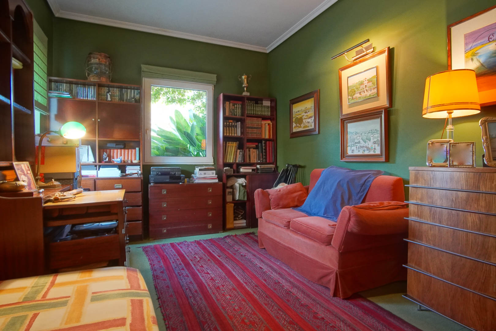 Despacho con paredes verdes y mobiliario de madera clásica. Cuenta con estanterías, escritorio, sofá rojo y luz natural con vistas a la vegetación exterior.