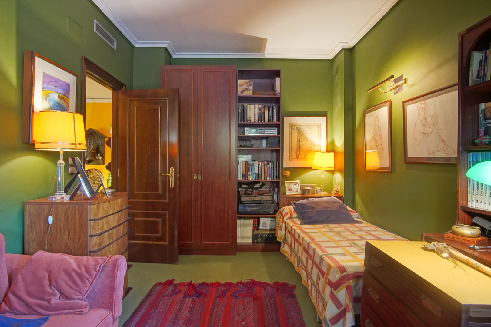 Dormitorio con paredes verdes, mobiliario de madera clásica, cama individual y estantería integrada. Incluye iluminación ambiental cálida y rejilla de aire acondicionado.