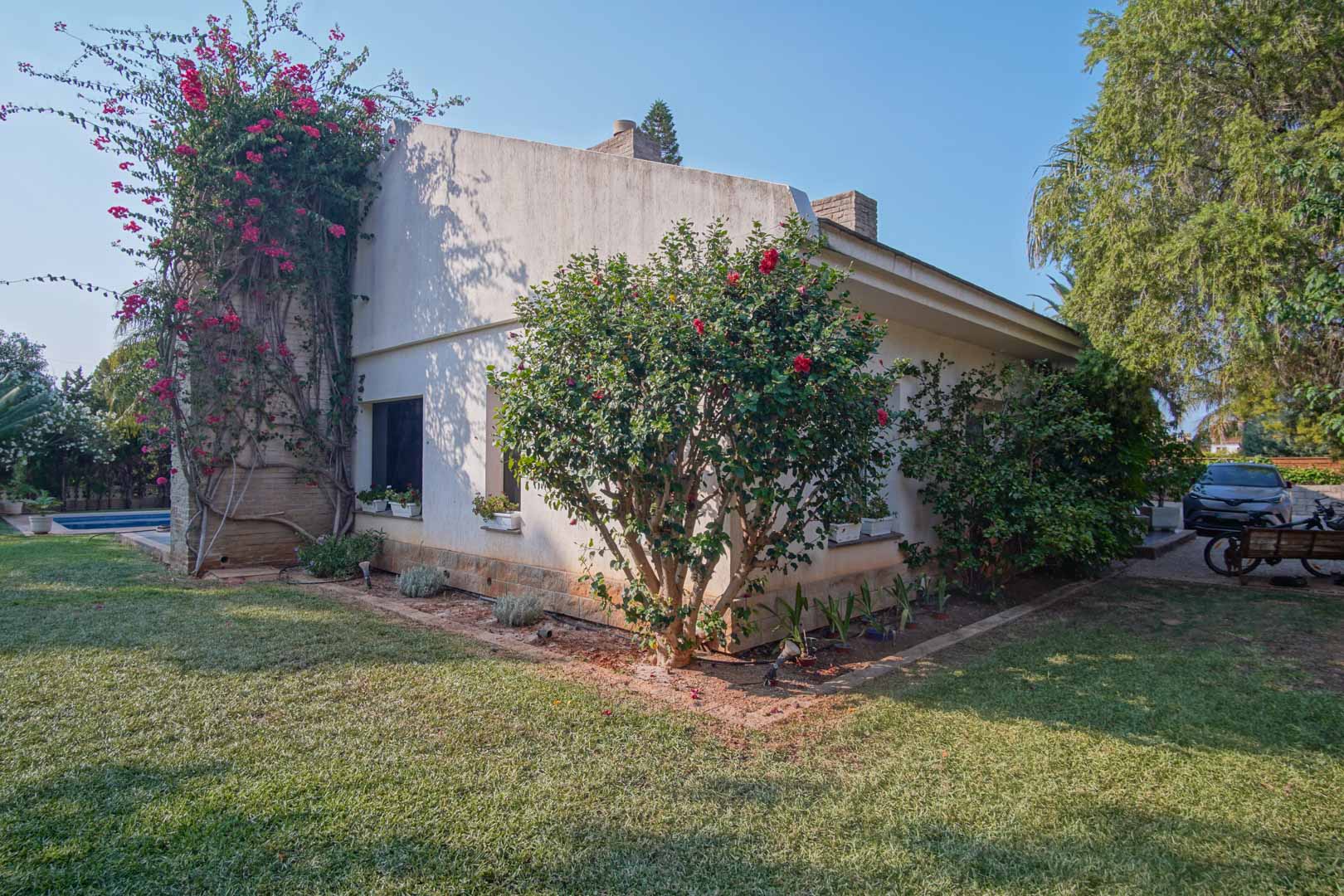 Fachada lateral de la villa con jardín de césped natural, buganvilla trepadora sobre muro de piedra y arbustos mediterráneos bajo un cielo despejado.
