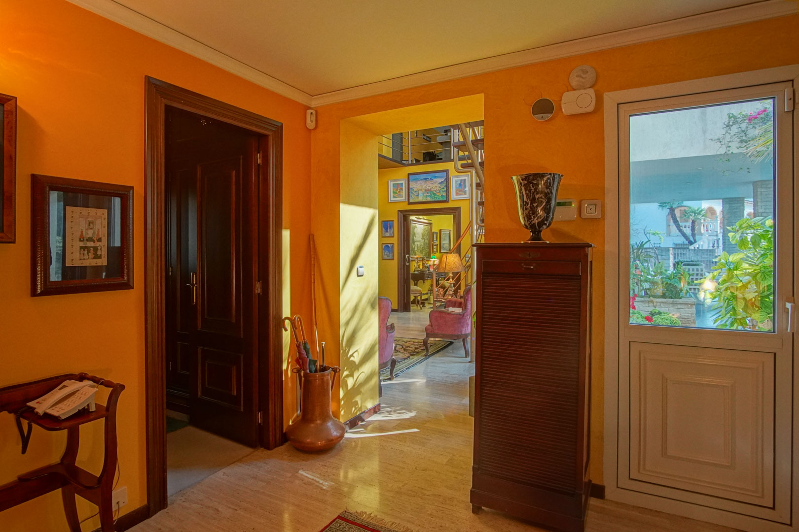 Hall de entrada con suelos de mármol, paredes en tonos cálidos y carpintería de madera noble. Ofrece acceso al salón y recibe abundante luz natural del exterior.