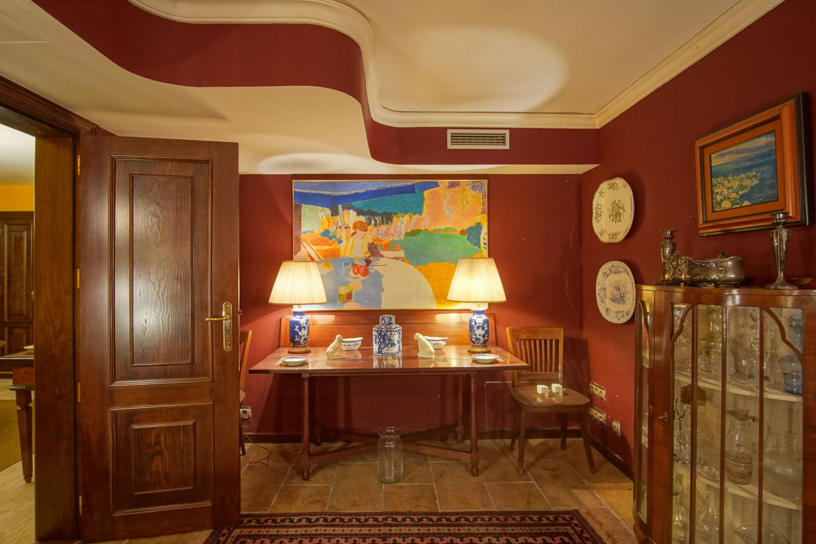 Rincón decorativo con paredes granate, techo de diseño ondulado y mobiliario de madera clásica. Incluye una vitrina acristalada y mesa auxiliar con lámparas cerámicas sobre suelo de piedra.