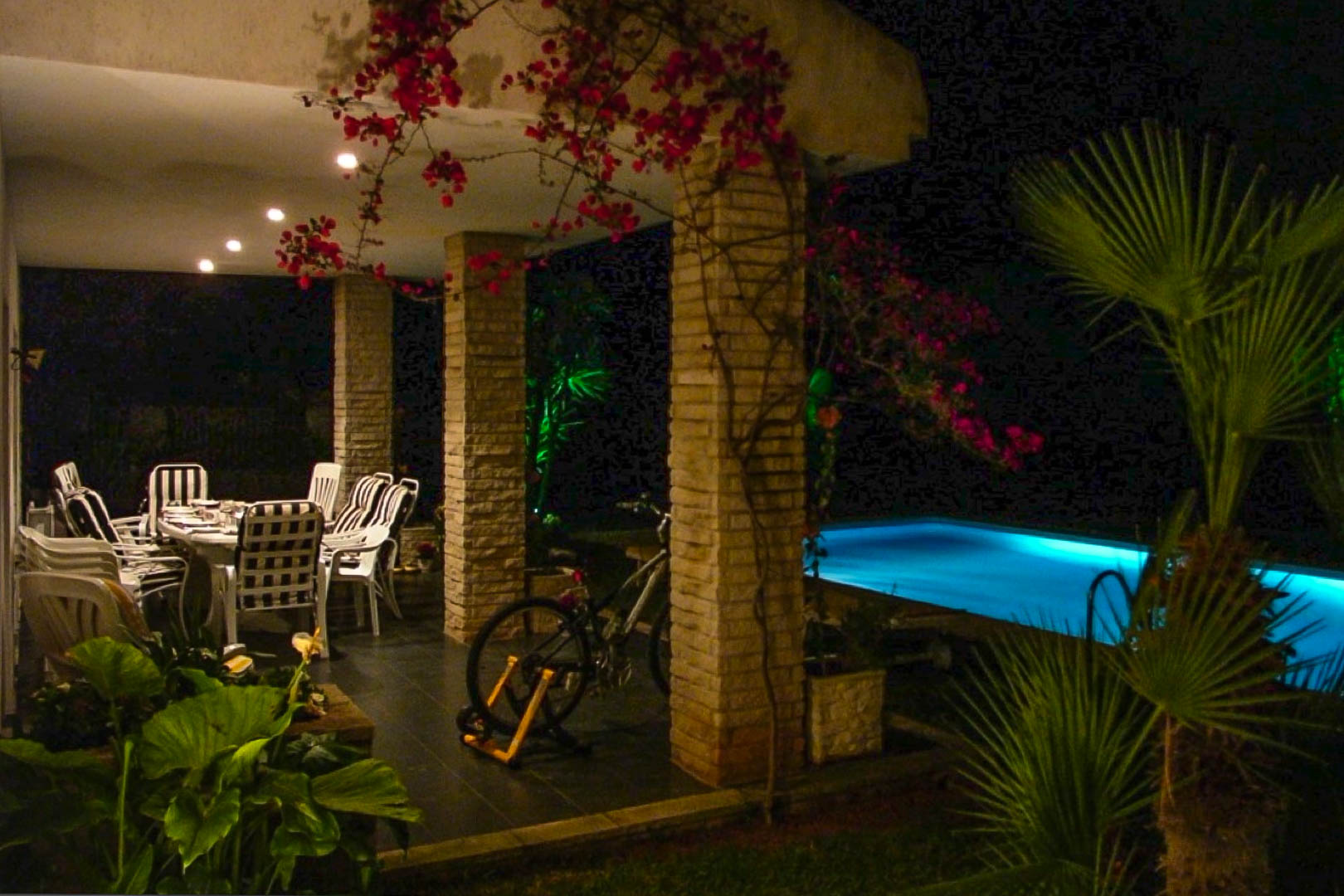 Terraza cubierta con columnas de ladrillo visto, comedor exterior y buganvillas, situada junto a una piscina iluminada en un entorno nocturno privado.