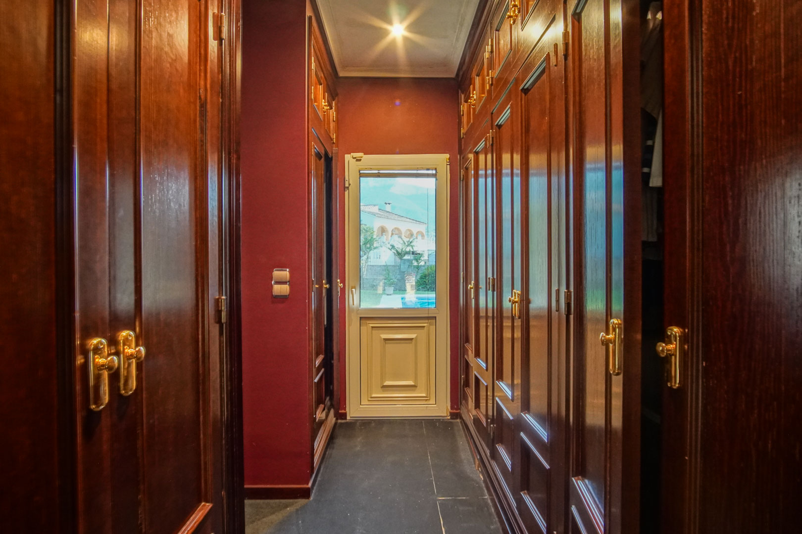 Vestidor con armarios empotrados de madera oscura y herrajes dorados, paredes granates y puerta acristalada con vistas al jardín.