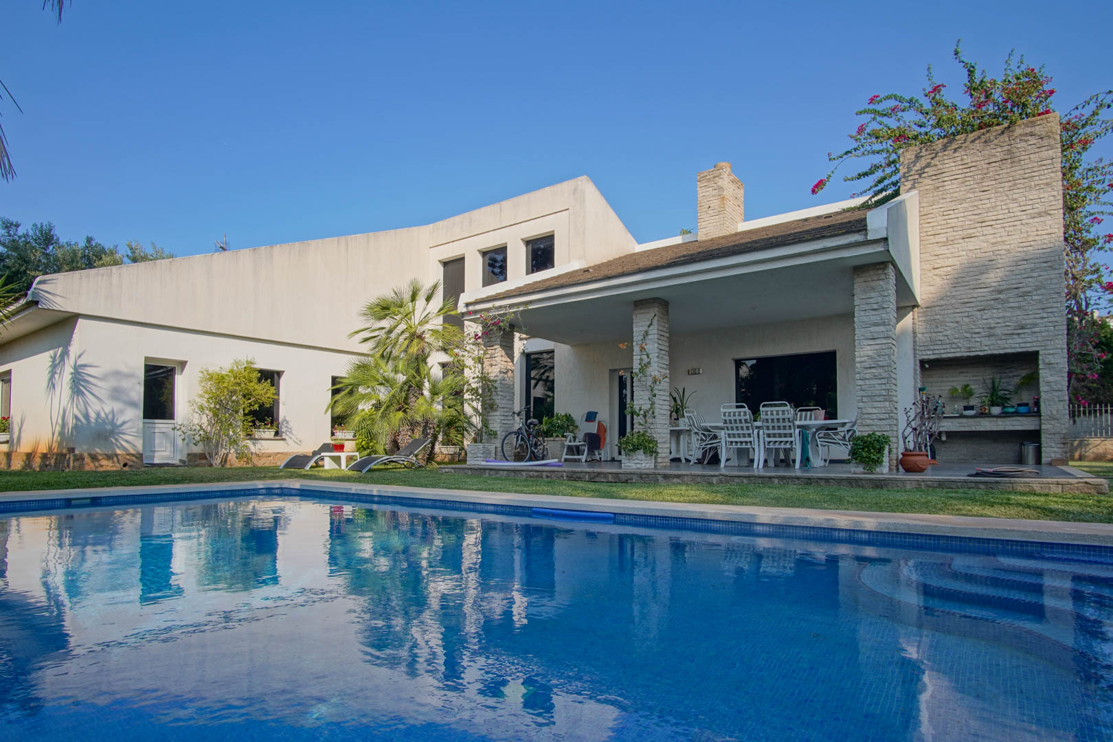 Vista exterior de la piscina privada y terraza cubierta con pilares de piedra, zona de comedor al aire libre y barbacoa integrada en un jardín mediterráneo.