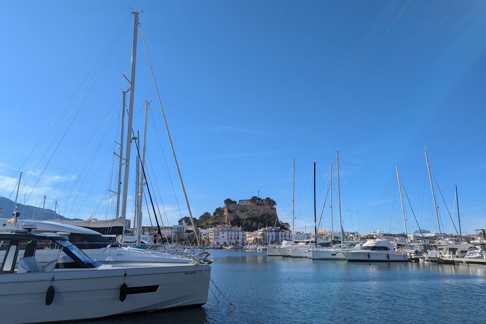 Vista panorámica del puerto deportivo de Dénia con embarcaciones de recreo y el histórico castillo presidiendo el paisaje urbano bajo un cielo despejado.