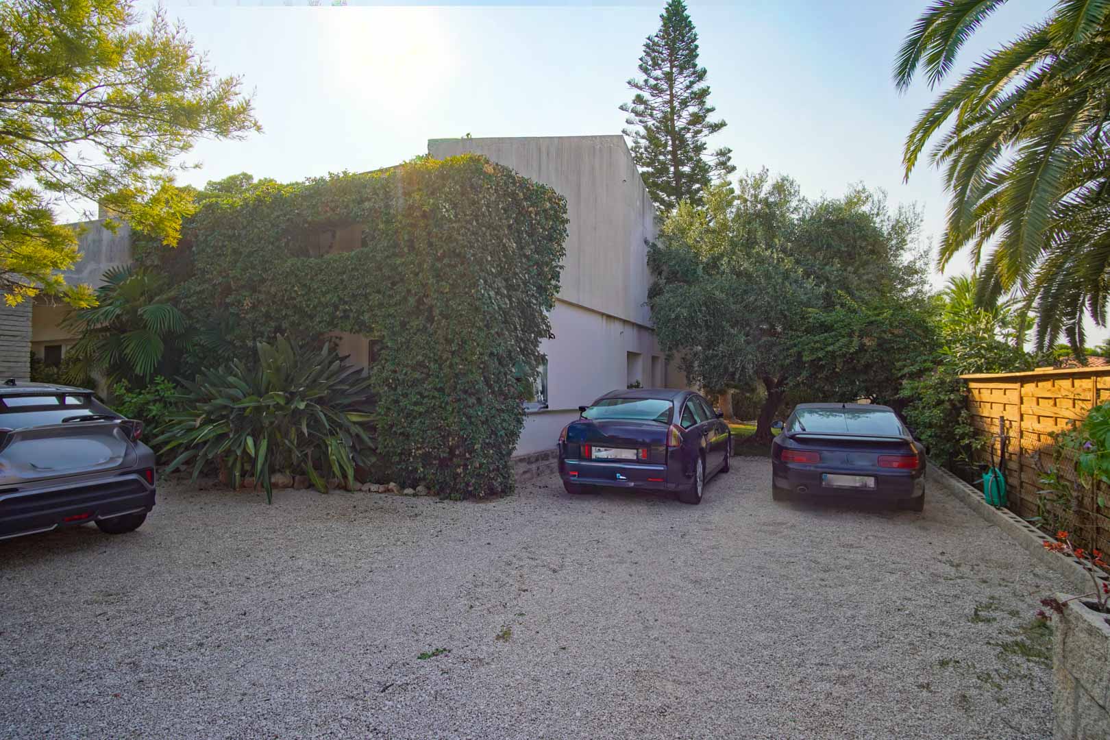 Zona de aparcamiento exterior con superficie de grava, rodeada de vegetación mediterránea madura y vallado perimetral de madera. Espacio con capacidad para varios vehículos.