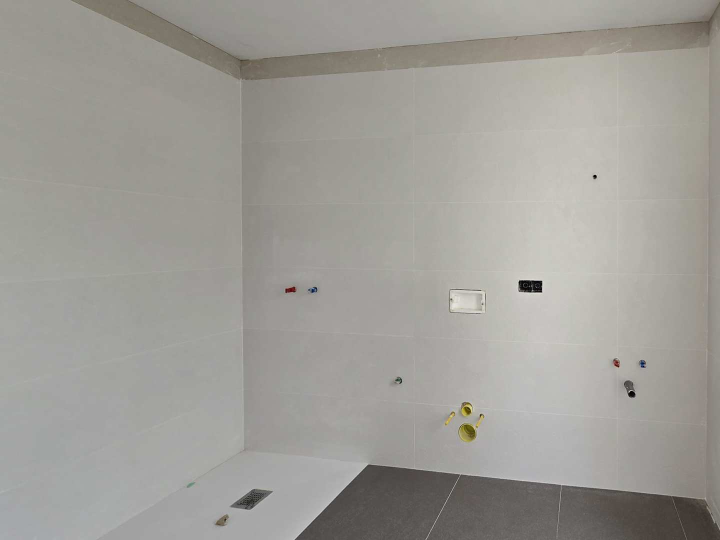 Baño en fase de construcción con revestimiento cerámico blanco, plato de ducha integrado a nivel del suelo y tomas de fontanería preparadas para los sanitarios.