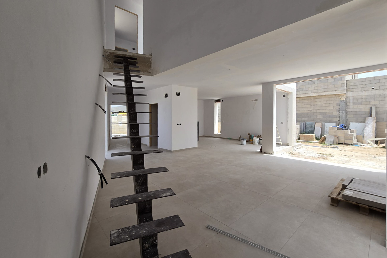 Interior diáfano en construcción con escalera metálica de diseño industrial, suelos de gres de gran formato y techos de doble altura que aportan amplitud.
