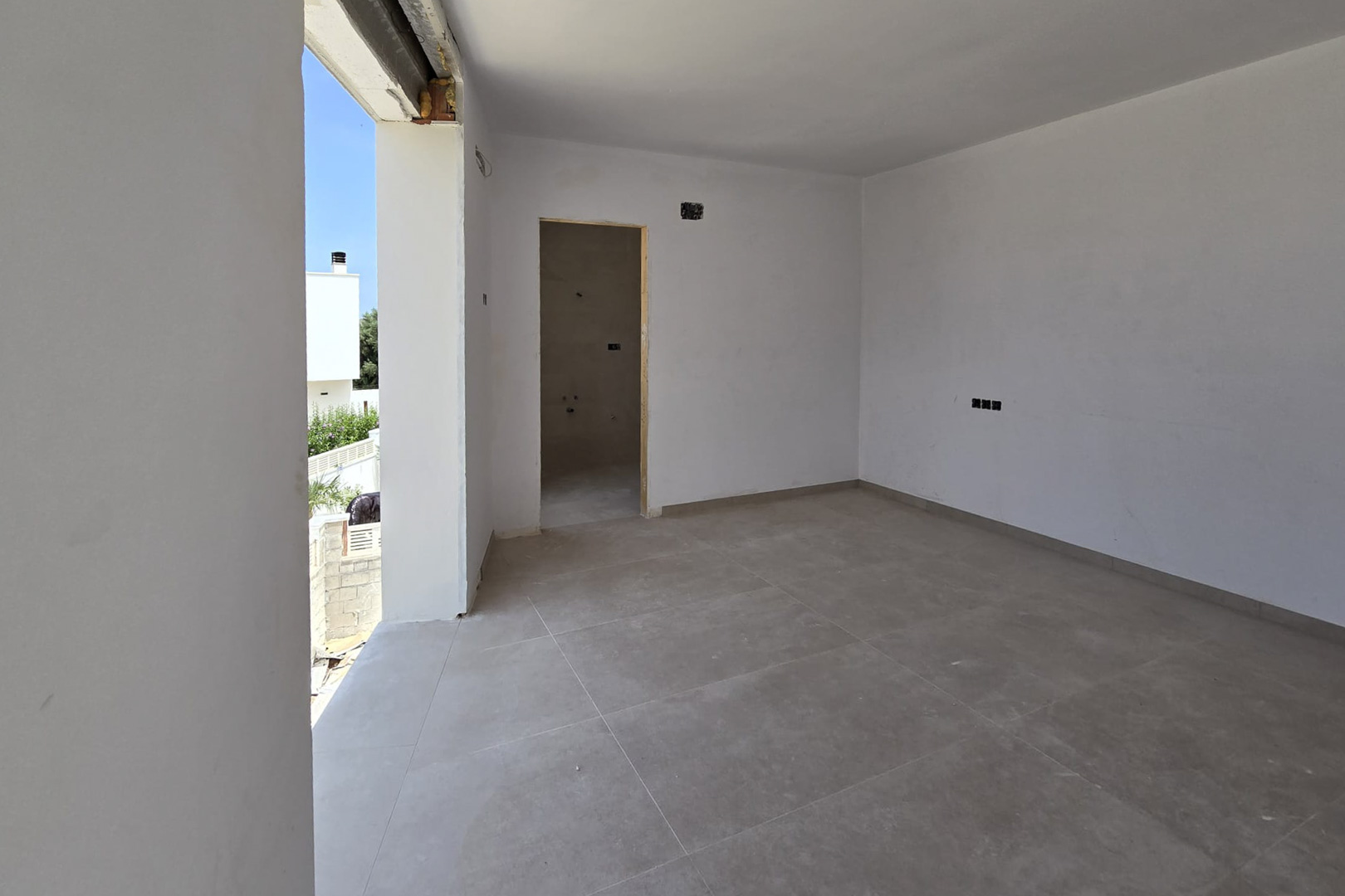 Interior en construcción con suelos de gres gris de gran formato, paredes blancas y acceso directo al exterior. Se observa el hueco para un baño en suite y tomas eléctricas.