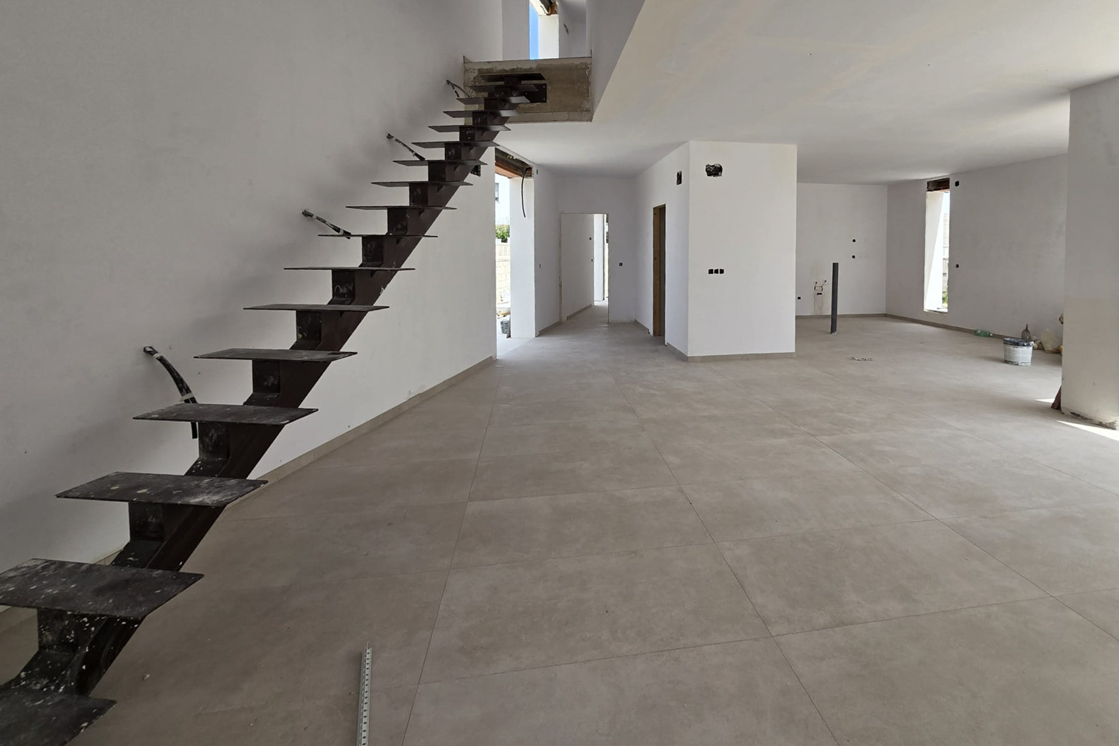 Planta baja diáfana en construcción con suelo porcelánico gris de gran formato y una escalera volada de estructura metálica que conecta con el nivel superior.