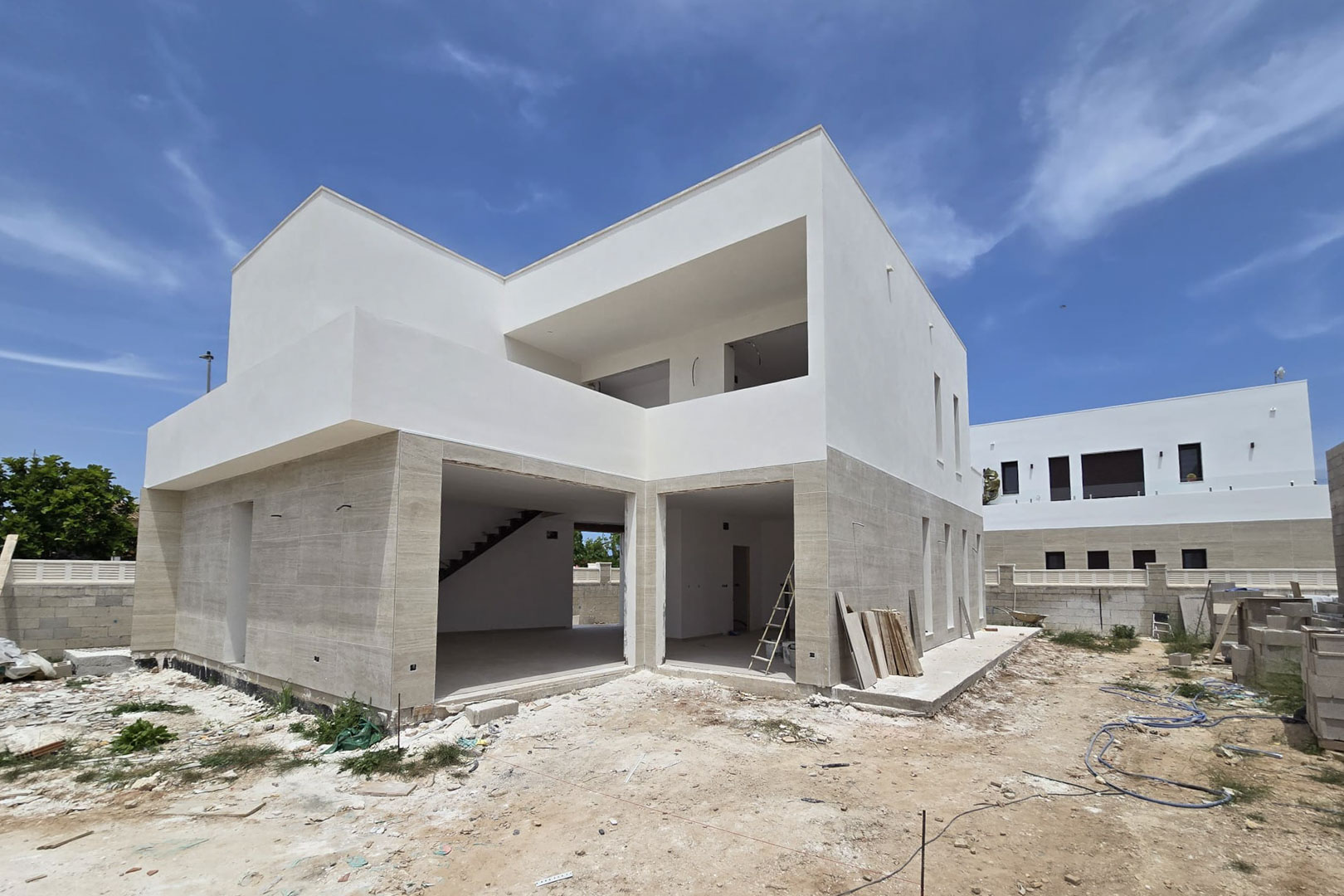 Vista exterior de la villa en construcción con fachada de piedra en planta baja y volúmenes blancos superiores, destacando los amplios huecos para ventanales.