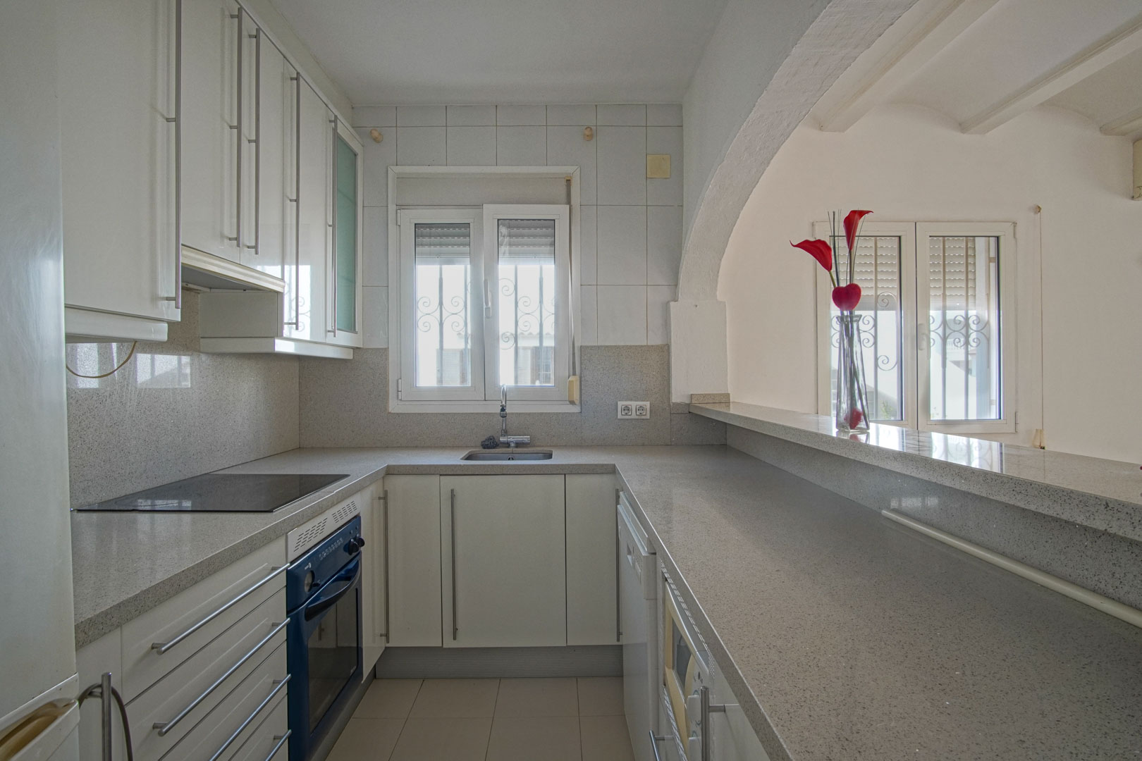 Cocina semiabierta con mobiliario blanco, encimera de granito gris y barra americana que conecta con el salón bajo un arco decorativo.