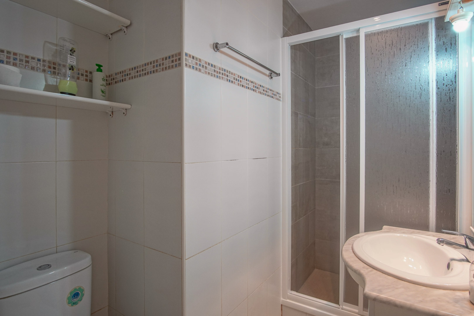 Cuarto de baño con paredes alicatadas en blanco y cenefa decorativa, equipado con ducha con mampara corredera y lavabo encastrado en encimera de mármol.