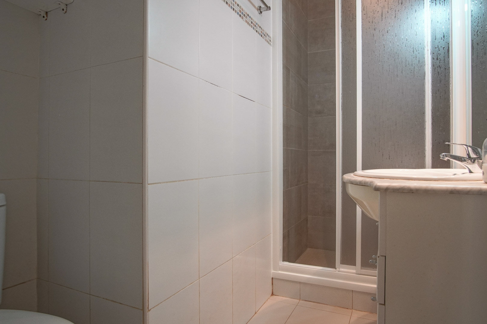 Cuarto de baño funcional con paredes alicatadas en blanco, cabina de ducha con mampara y lavabo encastrado sobre encimera de mármol.