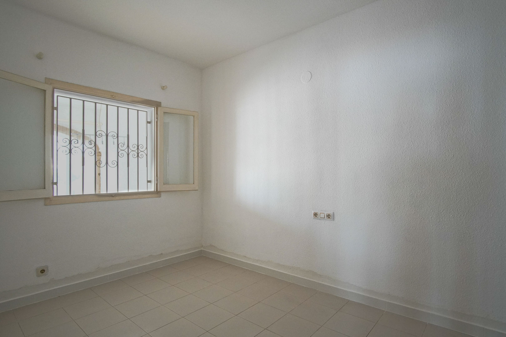 Dormitorio luminoso con paredes blancas texturizadas, suelos de gres claro y ventana exterior con rejas de seguridad decorativas.