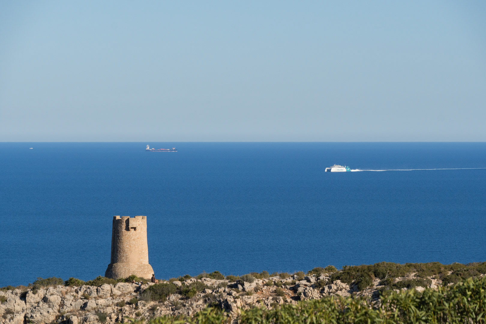 Vista de la histórica Torre del Gerro en los acantilados de Dénia, con el mar Mediterráneo y un ferry de Baleària navegando en el horizonte.